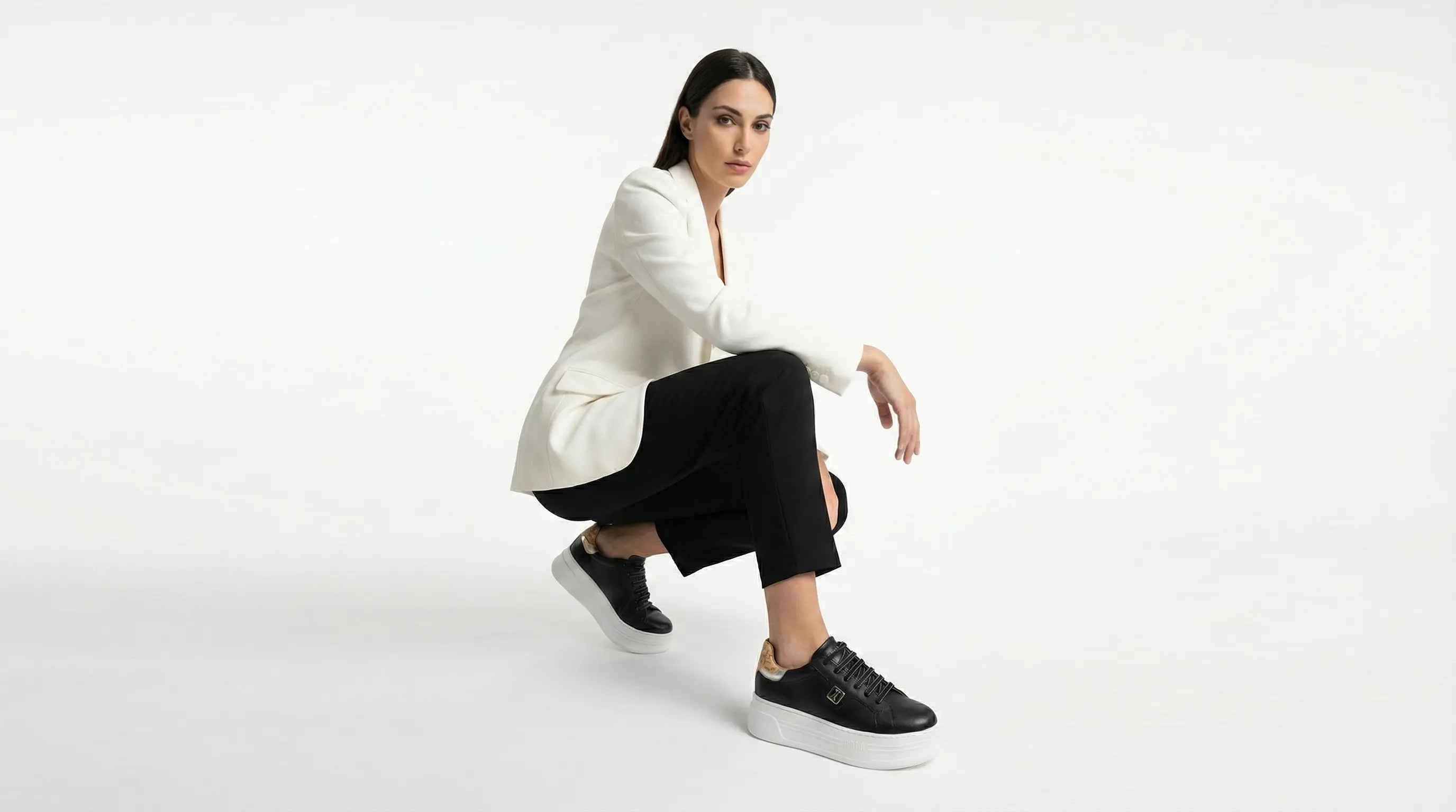 Woman sitting on a white background wearing a white blazer, black pants, and black sneakers.