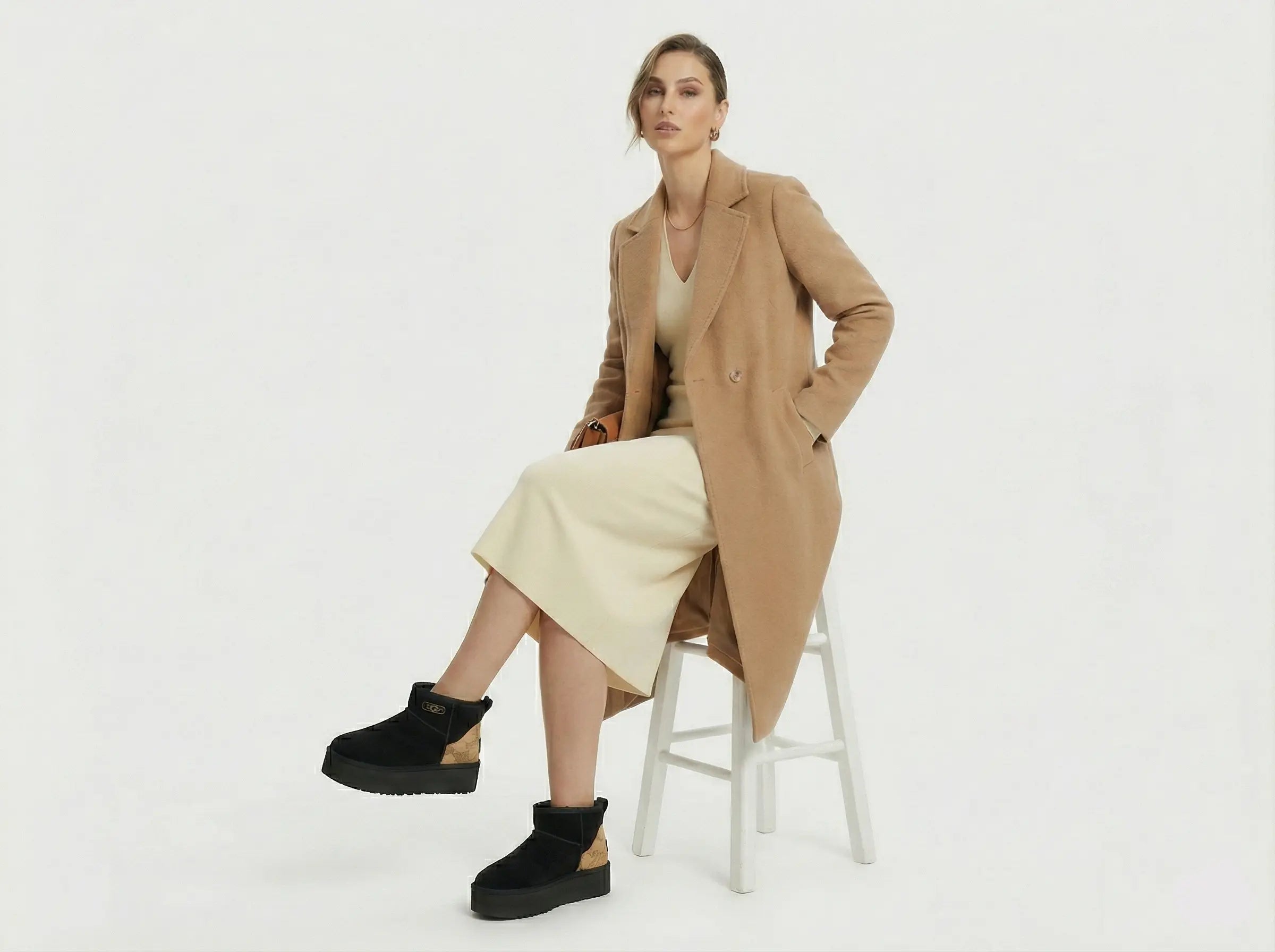 Woman wearing a beige coat and black platform shoes sitting on a white stool against a white background