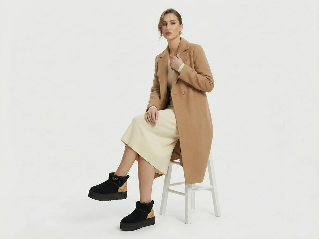 Woman wearing a tan coat and black platform shoes sitting on a white stool against a white background