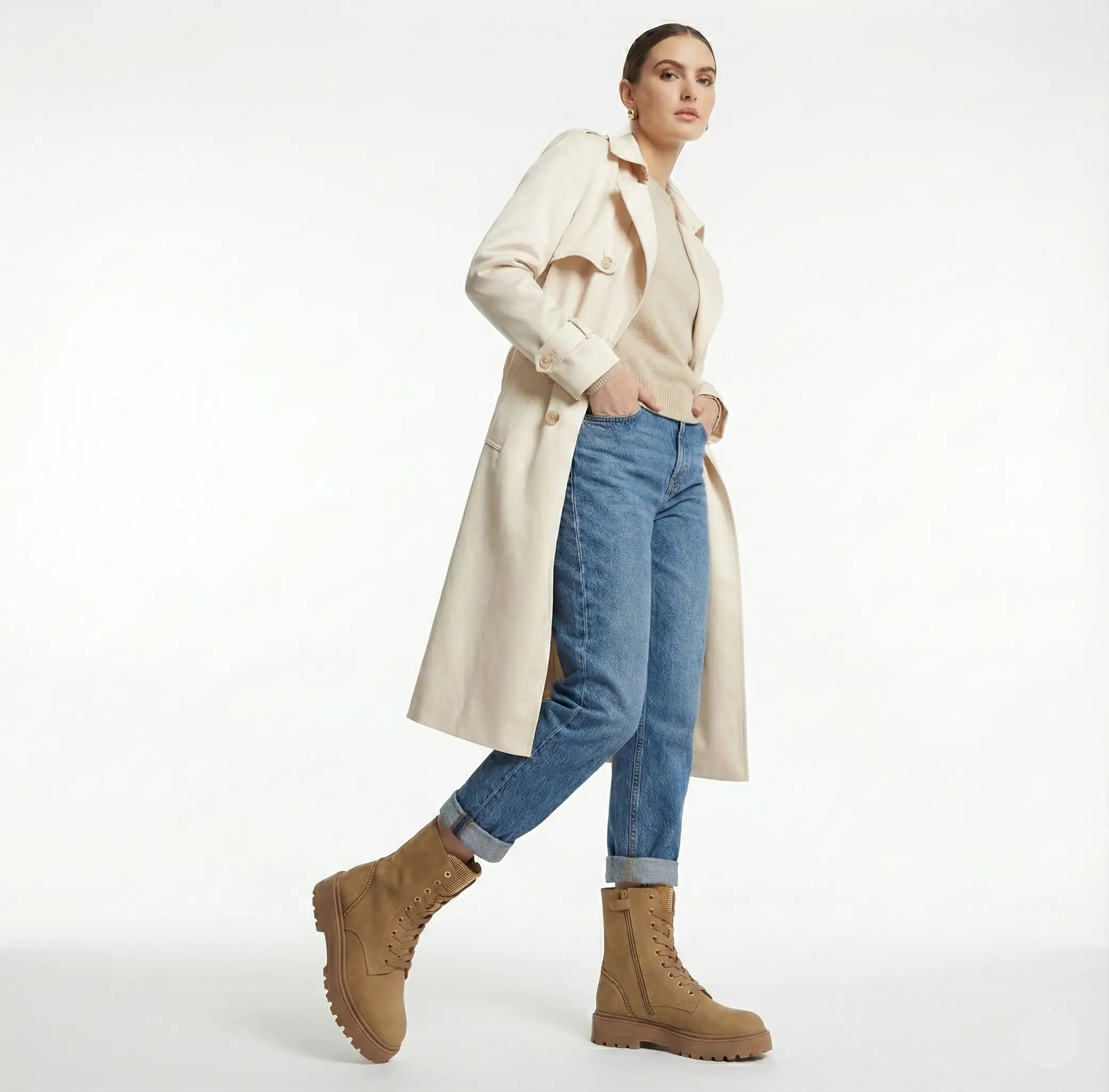 Woman wearing a beige trench coat, blue jeans, and brown boots on a white background