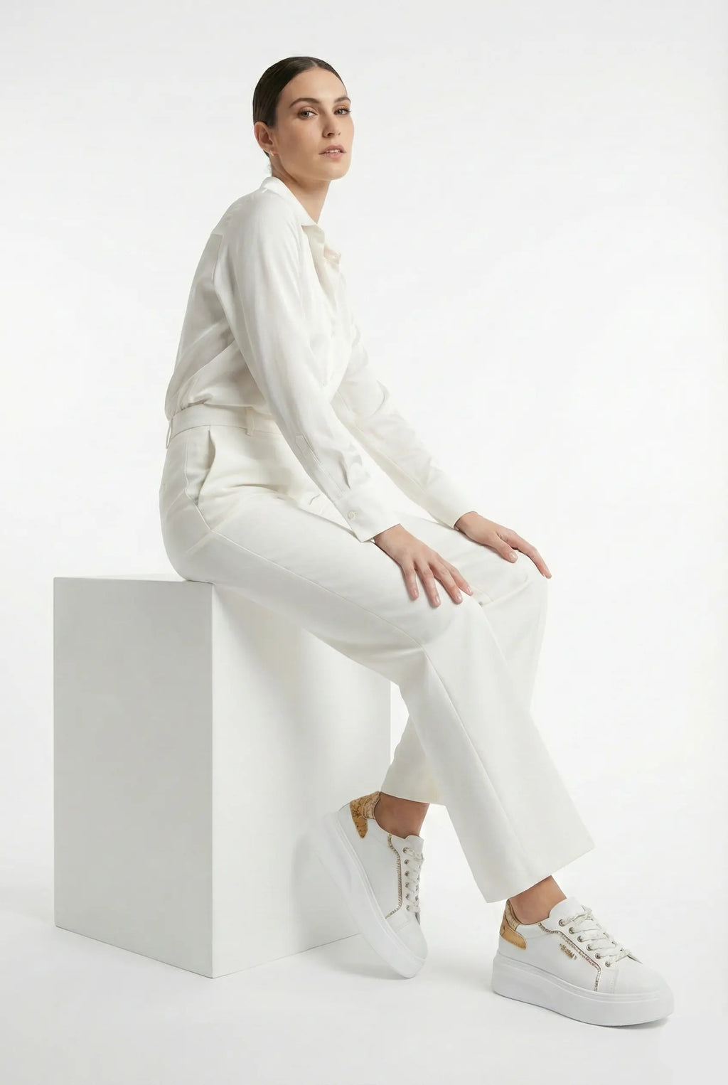 Person wearing a white outfit sitting on a white cube against a white background